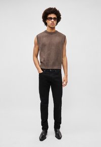 Suéter marrón sin mangas con un escote acanalado, combinado con jeans negros ajustados y zapatos negros elegantes, de pie frente a un fondo sencillo.