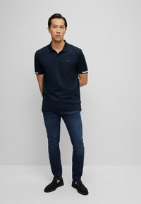 Tamsiai mėlynas polo marškinėliai su kontrastinėmis juostelėmis ant rankovių, logotipas ant krūtinės, derinami su tamsiai mėlynomis džinsinėmis kelnėmis ir tamsiais šlepetėmis.