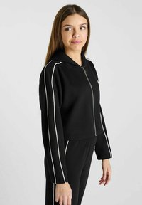 Zwarte vrouwelijke cropped hoodie met witte piping langs de mouwen, volledig rits voorkant en een capuchon. Heeft een ontspannen pasvorm en een zachte stoffen textuur.