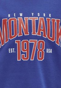 Blauwe katoenen t-shirt met rode tekst: "NEW YORK MONTAUK 1978 EST. USA." Vet, blokletters met een soepel oppervlak.