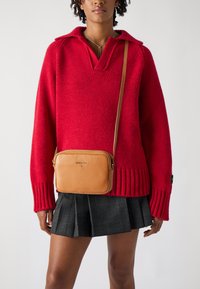 Maglione rosso lavorato a maglia con scollo a V e polsini a costine, abbinato a una borsa a tracolla in pelle beige e una gonna nera plissettata con motivo a scacchi.