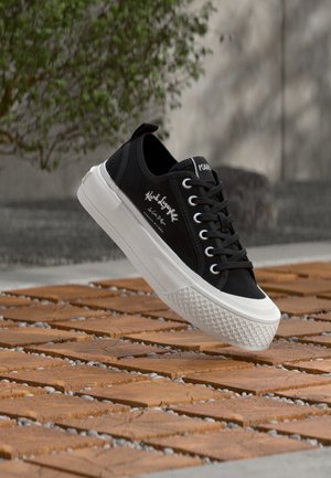 Baskets basses noires avec semelle blanche et lacets, logo en lettres cursives sur le côté, flottant au-dessus d'un sol en dalles en bois et galets en extérieur.