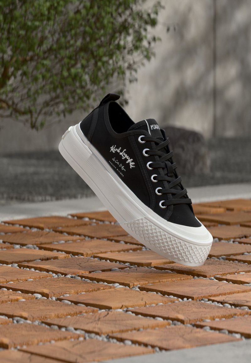 Μαύρο χαμηλό sneaker με λευκή σόλα και κορδόνια, καλλιγραφικό λογότυπο στο πλάι, που αιωρείται πάνω από ξύλινο πλακάκι και πετρώδη έδαφος σε εξωτερικό χώρο.