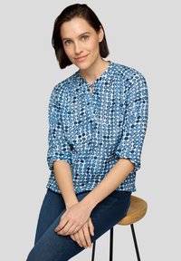 Blaues Hemd mit Knöpfen und einem kreisförmigen Muster in verschiedenen Farbtönen. Lange Ärmel hochgekrempelt. Glatte Textur. Getragen zu dunklen Jeans.