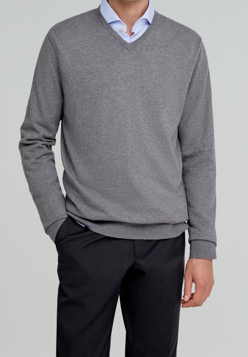 Homme portant un pull gris à col en V par-dessus une chemise bleu clair à col avec un pantalon noir, debout avec une main dans la poche devant un fond uni.