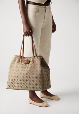 Beige tote bag with brown patterned detail and leather accents. It features long handles and a logo clasp, carried alongside cream pants and flats.