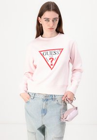 Sweat-shirt rose clair avec un logo triangulaire rouge comportant le texte "GUESS". Associé à un jean bleu clair ample et un petit sac à main rose.