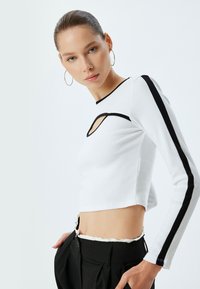 Witte longsleeve top met ribbelstructuur, met een uitsnijding op de borst en zwarte accentstrepen langs de mouwen. Gecombineerd met zwarte broek.