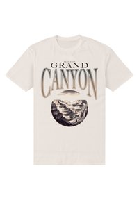 Cremefarvet t-shirt med et rundt grafisk billede af Grand Canyon, med sort tekst ovenfor og nedenfor, der forestiller bjerglandskaber.