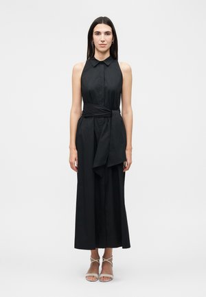 Kobieta stojąca, ubrana w czarną midi sukienkę bez rękawów z kołnierzykiem, przewiązaną w talii oraz beżowe sandały na paski na białym tle.