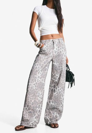 Femme portant un t-shirt court blanc noué, pantalon large gris à imprimé léopard, bracelets, tenant un sac à main noir, debout avec une main dans la poche.