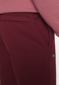 Bordové tepláky s hladkou texturou, s kapsou na zadní straně a malým černým logem Under Armour na boku.