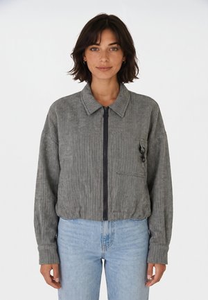 Femme aux cheveux courts et foncés portant une veste grise zippée et un jean bleu clair, debout devant un fond blanc uni.
