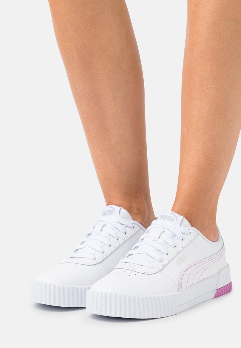 Jambes portant des baskets blanches Puma avec des touches roses, des semelles épaisses à rainures et des lacets blancs sur un fond clair uni.