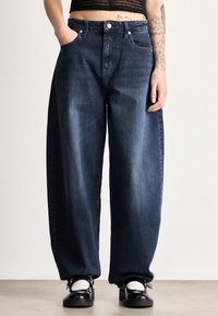 Tmavomodré široké rifle z denimu s vysokým pásom, vybavené prednými vreckami a minimálnym ošúchaním. Doplnene čierno-bielymi topánkami.