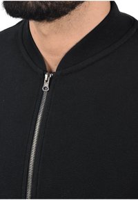 Schwarze Bomberjacke mit einem gerippten Kragen und einem silbernen Reißverschluss, gefertigt aus einem strukturierten Stoff, der für eine figurbetonte Passform sorgt.