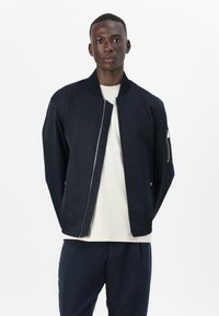 Veste de bombardier marine avec fermeture éclair, col et poignets côtelés. Comprend une poche latérale avec fermeture éclair. Portée par-dessus une chemise de couleur claire.