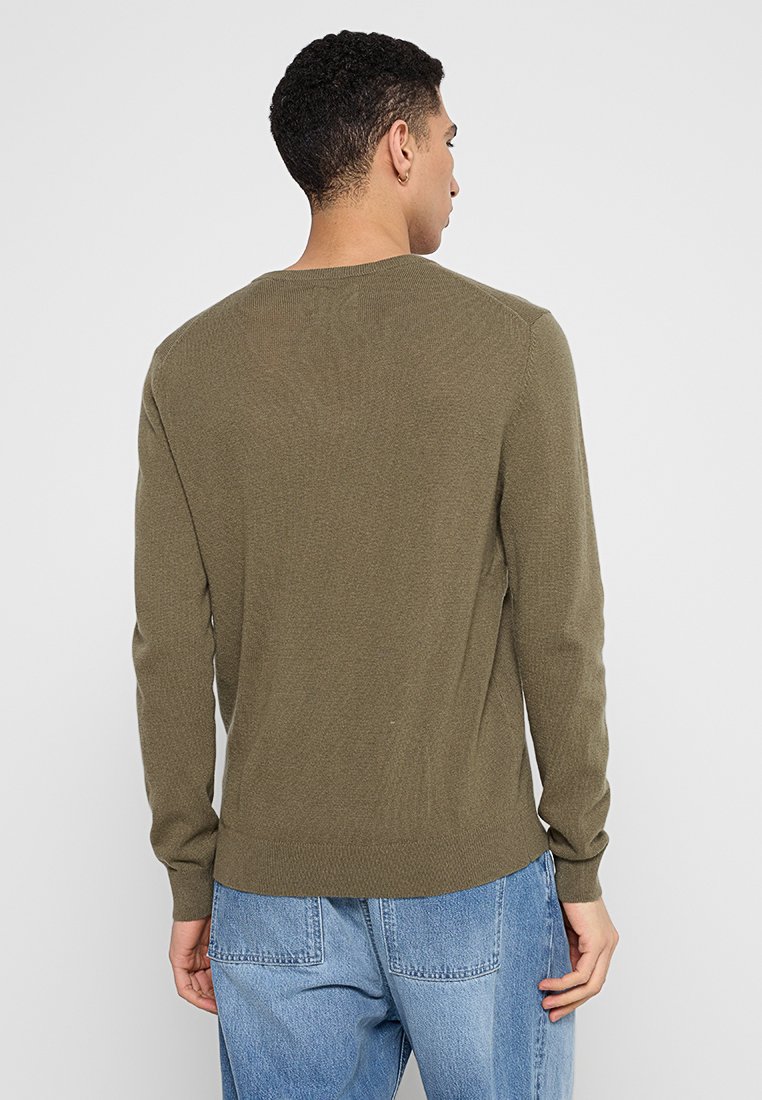 Olivgrön stickad tröja med långa ärmar, rundad halsringning och ribbad nederkant. Buren över blå denimbyxor, som visar en slät textur och figurnära design.