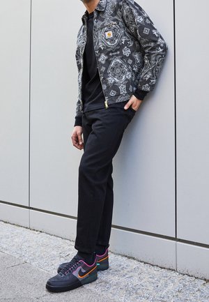 Person, der læner sig op ad en grå væg iført sorte bukser, sort-hvid mønstret jakke og sorte sneakers med orange og pink accenter.