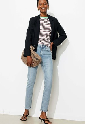 Femme souriante en chemise rayée, blazer noir, jean bleu clair et sandales noires à talons tenant un sac à main beige texturé contre un mur blanc.