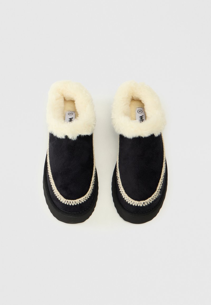 Pantofole nere in suede con fodera in ecopelliccia crema, cuciture a contrasto, forma arrotondata e suola spessa nera. Etichetta della taglia visibile all'interno.