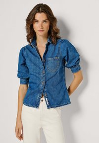 Młoda kobieta o falujących brązowych włosach, ubrana w niebieską koszulę jeansową z podwiniętymi rękawami i białe spodnie, stojąca na jednolitym tle.