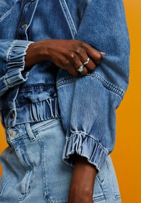 Denimjacka med elastiska ärmslut och midja. Ljusblått tyg med lätt slitna mönster. Hand prydd med ringar, mot en orange bakgrund.