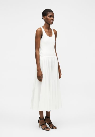 Γυναίκα που φοράει λευκό αμάνικο τοπ και λευκή φούστα μέσης μήκους που ρέει, με μαύρα ψηλοτάκουνα σανδάλια με λουράκια, σε απλό φόντο.