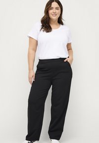 Zwarte wijde broek met een gladde textuur en een elastische tailleband, gecombineerd met een eenvoudig wit T-shirt. Model staat met één hand in de zak.