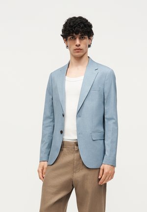 Νέος άνδρας με σγουρά μαλλιά που φοράει ένα ανοιχτό μπλε σακάκι, λευκό φανελάκι και μπεζ παντελόνι, στέκεται μπροστά σε ένα λευκό απλό φόντο.