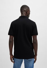 Schwarzes Polo-Shirt mit klassischem Kragen, kurzen Ärmeln und glatter Textur; kombiniert mit hellblauen Jeans. Rückansicht gezeigt.