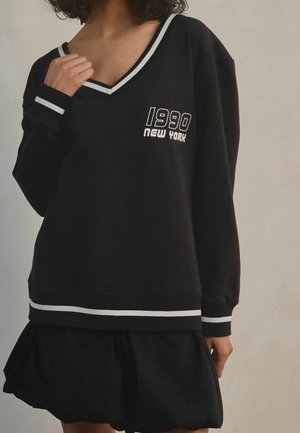 Personne portant un pull noir à col en V avec des rayures blanches et le texte « 1990 New York », associé à une jupe noire.