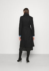 Zwarte, lange wollen jas met een ceintuur in de taille, hoge kraag en een rechte pasvorm. Getextureerde stof met een gladde afwerking, gelaagd over een donkere rok.