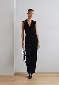 Salopette noire sans manches avec un décolleté en V profond, des accents d'épaules studded, et une taille cintrée, associée à une pochette métallique et des bottines.