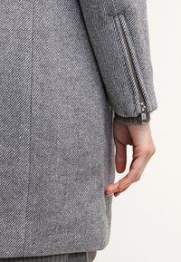 Šedý vlněný kabát s rybí kostí texturou, zipovými manžetami a střihovým střihem, ruka opřená po straně.