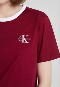 Osoba ve vínovém tričku s krátkým rukávem a bílým límečkem, na kterém je vyšitý logo "CK Calvin Klein Jeans" na hrudi.