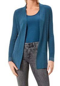 Teal färgad cardigan med ribbad textur och öppen front. Långa ärmar, lager på en matchande tanktop, parat med grå jeans.