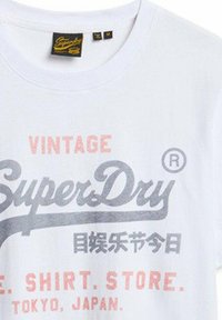 Camiseta de algodón blanca con la palabra "Vintage" en rosa, "SuperDry" en gris y texto adicional en varios colores; escote redondo, etiqueta visible.