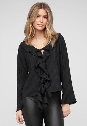 Ung kvinde med skulderlangt blondt hår iført en sort bluse med lange ærmer og flæser foran samt sorte læderbukser, stående.