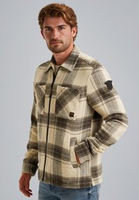 Geruite fleece jas in crèmelig bruin met een ritssluiting aan de voorkant, twee borstzakken en een logo patch op de mouw. Zachte textuur, relaxed fit.