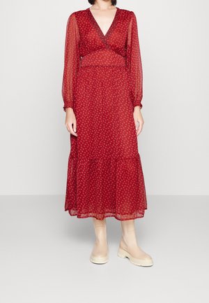 Frau trägt ein rotes Midi-Kleid mit langen Ärmeln und kleinen weißen Mustern sowie cremefarbene Stiefeletten und steht vor einem schlichten hellen Hintergrund.