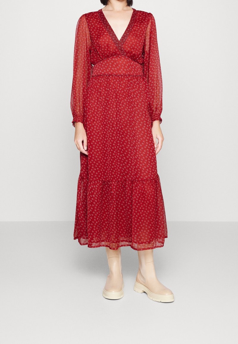 Femme portant une robe midi rouge à manches longues avec de petits motifs blancs et des bottines crème, debout devant un fond clair uni.