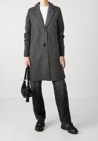 Osoba v tmavě šedém vlněném kabátu, černých džínách, černých teniskách, držící malou černou kabelek na pozadí jedné světle šedé barvy.