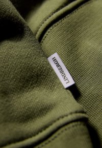 Olivgrön tyg med en texturerad yta, med en vit etikett som har "LINDENBERG" broderat på sig. Närbild av kläddetalj.