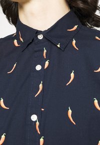 Camisa azul marino con botones, bordada por completo con chiles rojos y cuello clásico. Cuenta con pequeños botones blancos y una textura suave.