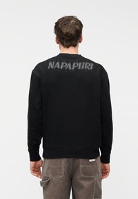 Άνδρας που φοράει μαύρο φούτερ με φθαρμένο κείμενο "NAPAPIJRI" στην πλάτη και καφέ παντελόνι εργασίας, γυρισμένος πλάτη σε απλό λευκό φόντο.