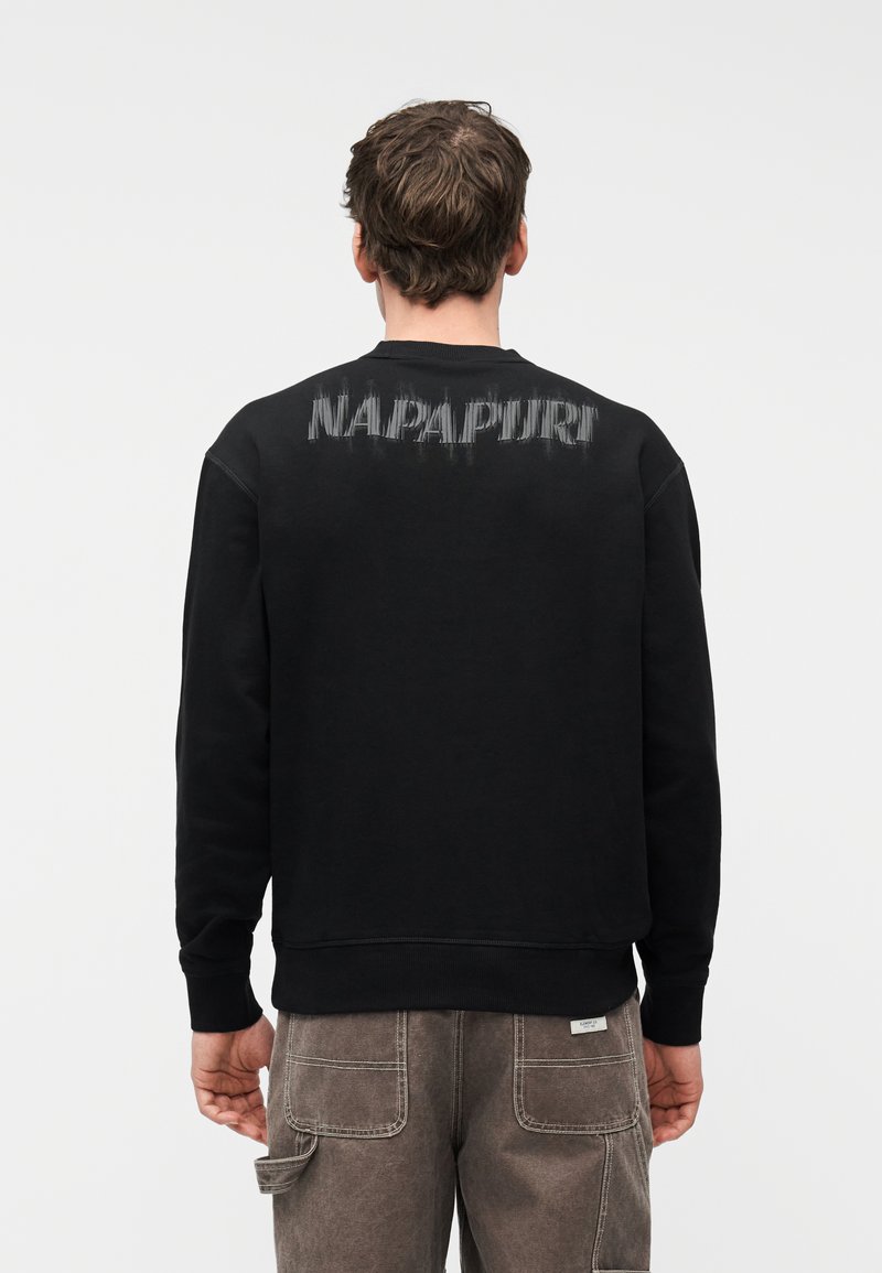 Άνδρας που φοράει μαύρο φούτερ με φθαρμένο κείμενο "NAPAPIJRI" στην πλάτη και καφέ παντελόνι εργασίας, γυρισμένος πλάτη σε απλό λευκό φόντο.