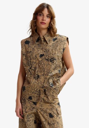 Donna che indossa una maglietta smanicata color sabbia e pantaloni abbinati con grande stampa floreale nera, in piedi con una mano in tasca su sfondo bianco.