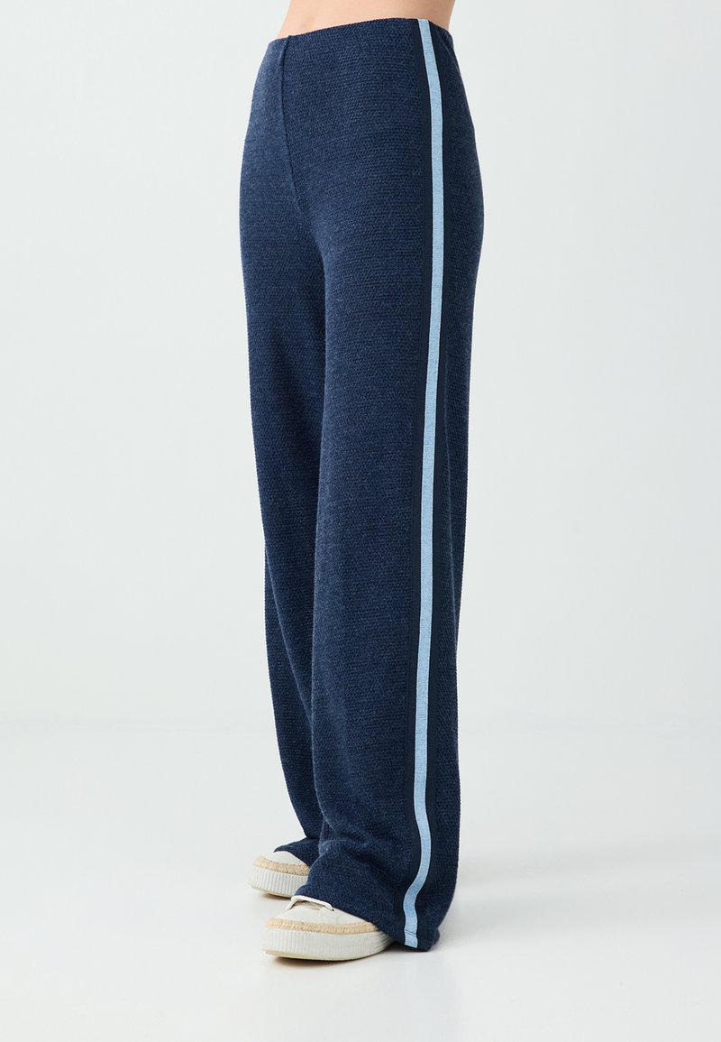 Pantalon large bleu marine avec des bandes latérales bleu clair porté avec des chaussures décontractées couleur crème.