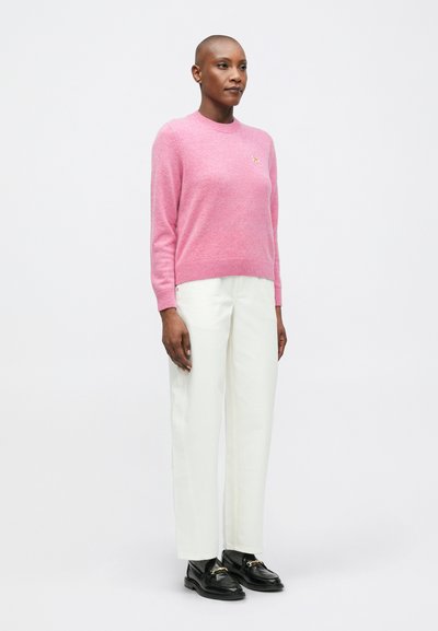 Osoba w różowym swetrze z długim rękawem, białych prostych spodniach, szarych skarpetkach i czarnych mokasynach stoi na tle jednolitego białego tła.